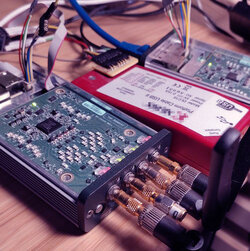 Two technical devices of the researchers are lying on a table surrounded by and connected with cables. On the left side is the USRP E310 from Ettus, on the right side is a device for FPGA debugging - the Chip-Scope from Xilinx.