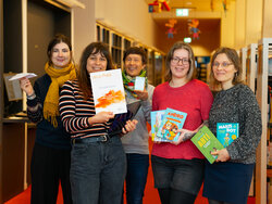 5 Frauen stehen in der Bibliothek. Eine hält einen Papierflieger, eine die Urkunde zum Le-La-Preis, eine den Pokal. die anderen beiden halten jeweils mehrere Kinderbücher zum Thema "Kinder und Roboter" in den Händen. Alle lächeln.
